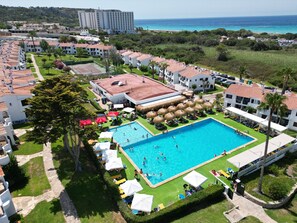 Un complexe hôtelier doté d’une piscine centrale entourée de transats et de parasols, d’un court de tennis et offrant une vue sur la mer.