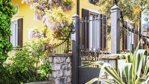 A yellow building with a stone staircase leading to a gated entrance, surrounded by greenery and a pergola with purple flowers.