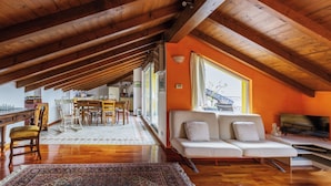 A spacious loft with a high ceiling, wooden beams, a cozy seating area with a sofa and TV, a dining area with chairs and a table, and a large window providing natural light.