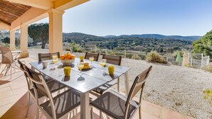 Un área de comedor al aire libre con una mesa dispuesta para cuatro personas, rodeada de sillas y con vistas a un paisaje pintoresco con colinas y una cerca.
