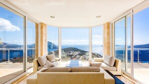 Een moderne kamer met een ruime bank en twee fauteuils, gericht op een panoramisch uitzicht over de zee door ramen van de vloer tot aan het plafond.