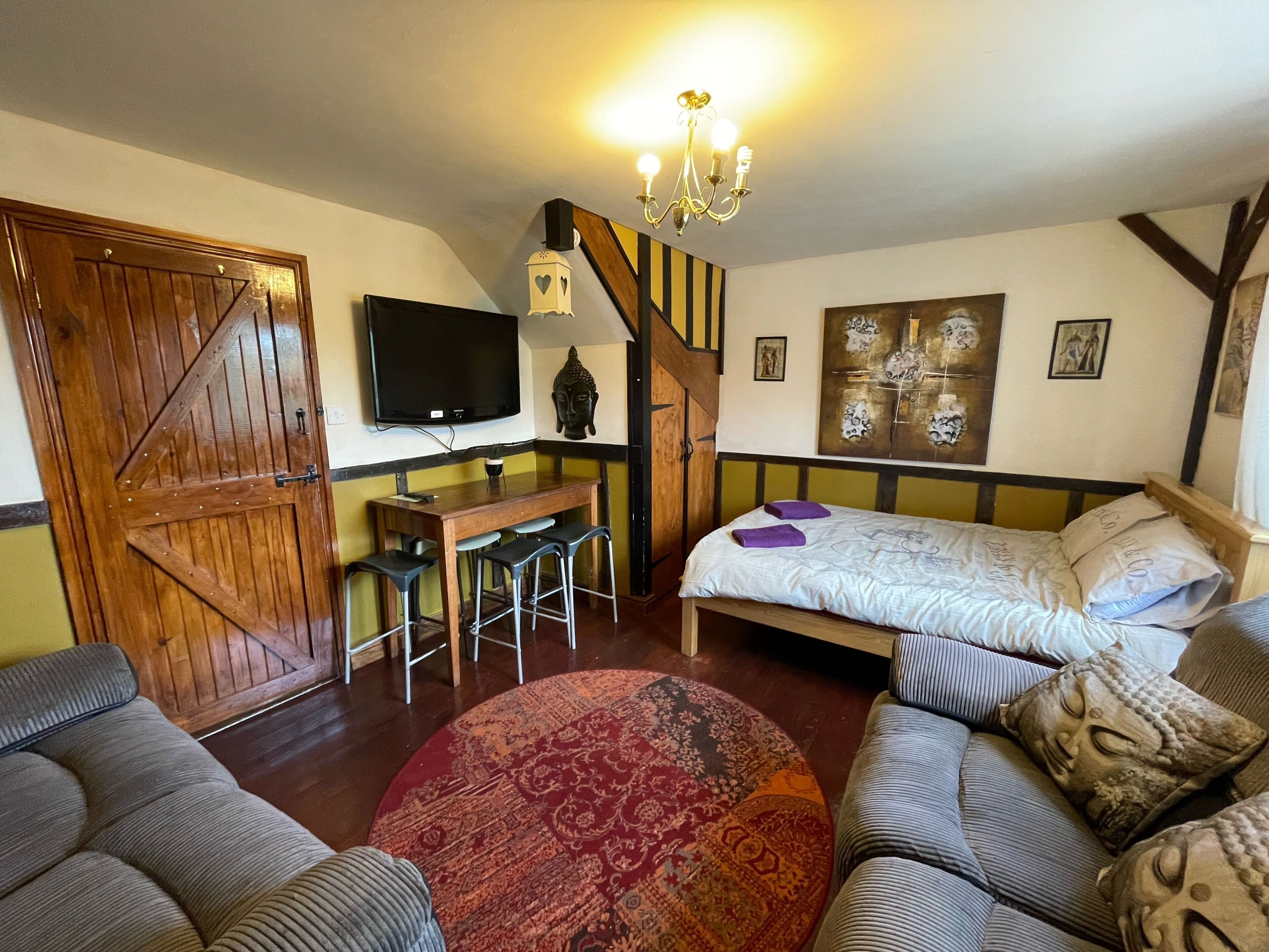 A cozy room with a bed, a sofa, a desk with two chairs, a TV, and a chandelier.