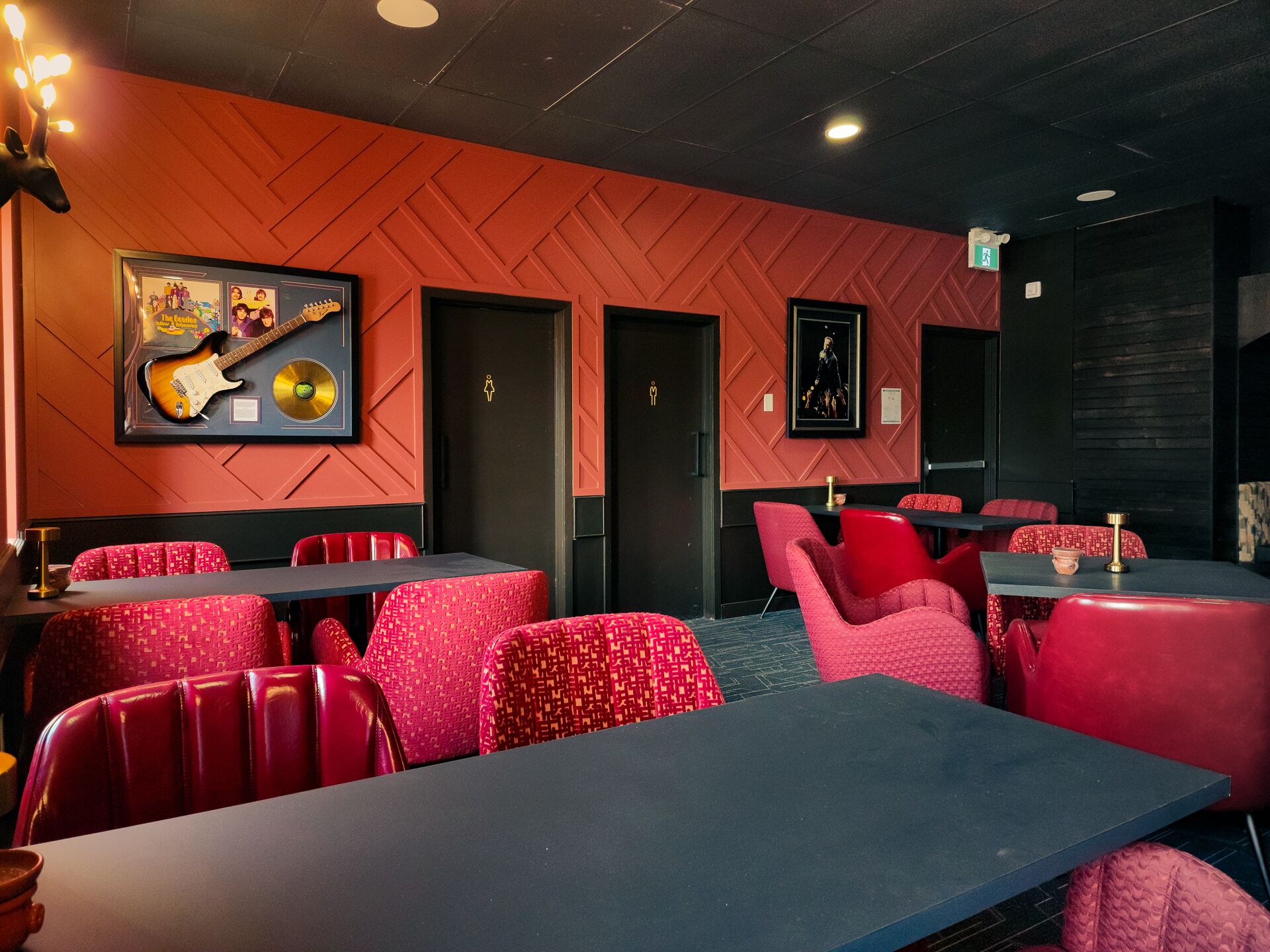 A restaurant with red upholstered booths and tables, a guitar and CD on the wall, and a black ceiling.