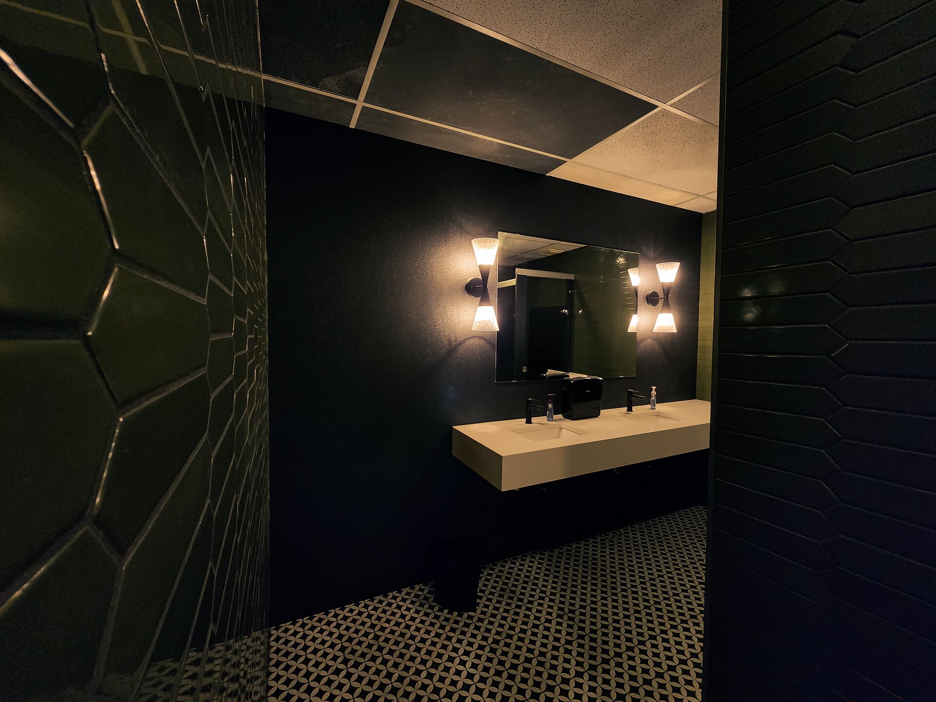 Un baño moderno con una pared oscura, un piso de diseño geométrico y un tocador blanco con un espejo y dos luces empotradas.