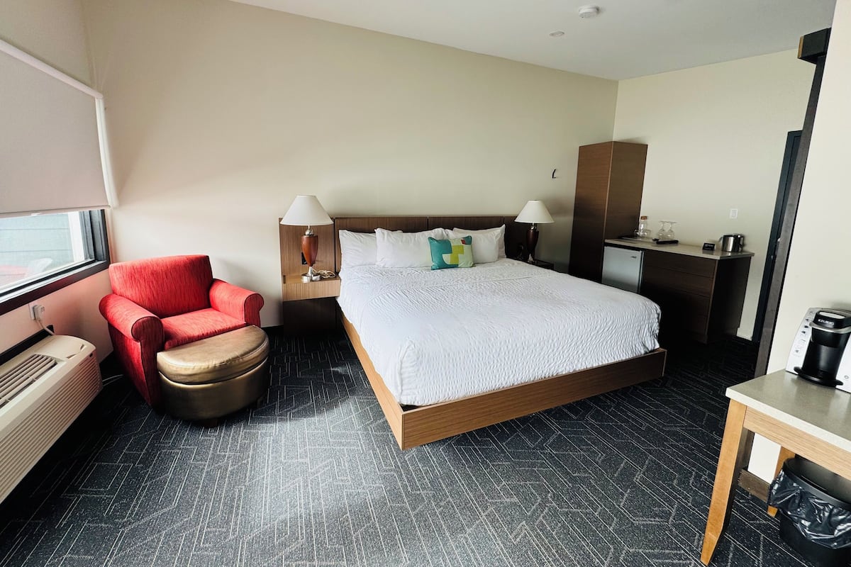 A hotel room with a bed, a red armchair, a desk with a coffee maker, and a minibar.