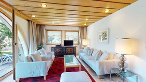 A spacious living room with a wooden ceiling, two white sofas, a glass coffee table, and a television on a cabinet. - Authentic semi-detached holiday home in a quiet location on the outskirts of Cel (Celerina/Schlarigna)