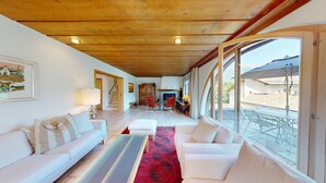 A spacious living room with a wooden ceiling, a fireplace, and a large window overlooking a patio. - Authentic semi-detached holiday home in a quiet location on the outskirts of Cel (Celerina/Schlarigna)