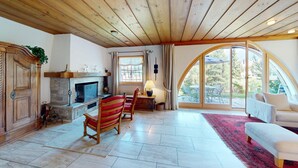 A spacious living room with a fireplace, a dining area with a table and chairs, and a large arched window offering a view of the outdoors. - Authentic semi-detached holiday home in a quiet location on the outskirts of Cel (Celerina/Schlarigna)