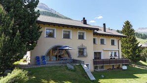 A two-story house with a patio, a covered outdoor seating area, and a mountain backdrop. - Authentic semi-detached holiday home in a quiet location on the outskirts of Cel (Celerina/Schlarigna)