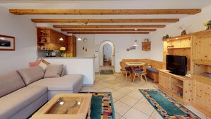 Ein gemütliches Wohnzimmer mit Sofa, Couchtisch, Holzbalken an der Decke, einer Küchenzeile mit Holzschränken und einem Fernseher.