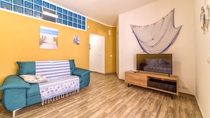Un soggiorno con un divano color petrolio, una coperta a righe, una TV su un mobile in legno e un'ampia finestra con infissi blu.