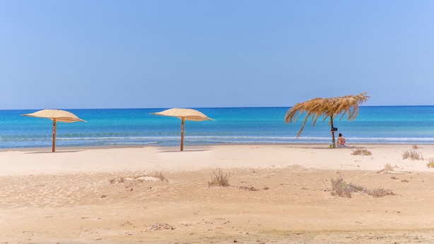 Una spiaggia con due ombrelloni di paglia e una persona seduta sulla sabbia.