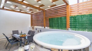 Patio esterno con idromassaggio, tavolo da pranzo, sedie e vista su un siepe verde.