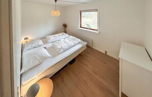 Een enkel bed met witte lakens, een klein nachtkastje, een hanglamp, een raam met zonwering en uitzicht op bomen.