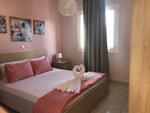 A bedroom with a bed, pillows, a nightstand, a lamp, a window with curtains, and wall art. - Omega Comfy Apartments (Apokoronas)