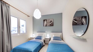 A room with two beds, each with a blue bedspread, a round mirror, and artwork on the wall. - Omega Comfy Apartments (Apokoronas)