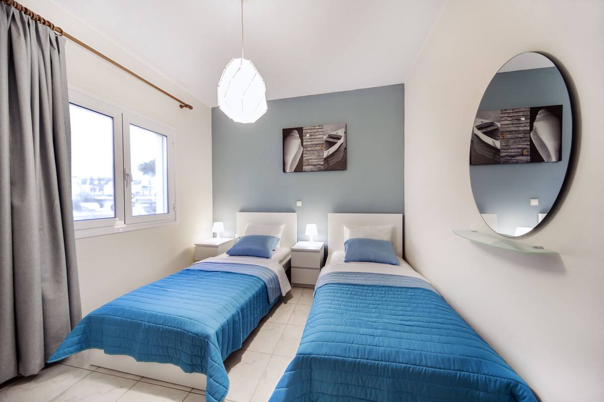 Ein Zimmer mit zwei Betten, jeweils mit blauer Bettdecke, einem runden Spiegel und Bildern an der Wand.