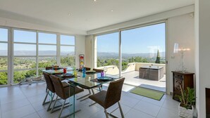 Un vaste espace de salle à manger avec une table et des chaises en verre, de grandes fenêtres donnant sur la campagne et une lampe moderne placée sur le côté.