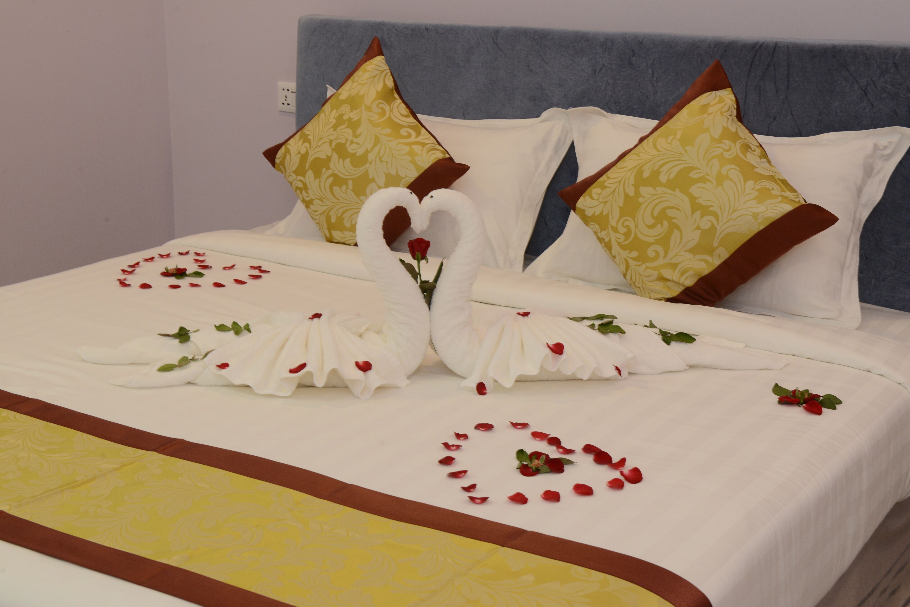 Una cama con colcha blanca y amarilla, dos almohadas con forma de cisne y arreglos de pétalos de rosa en forma de corazón.