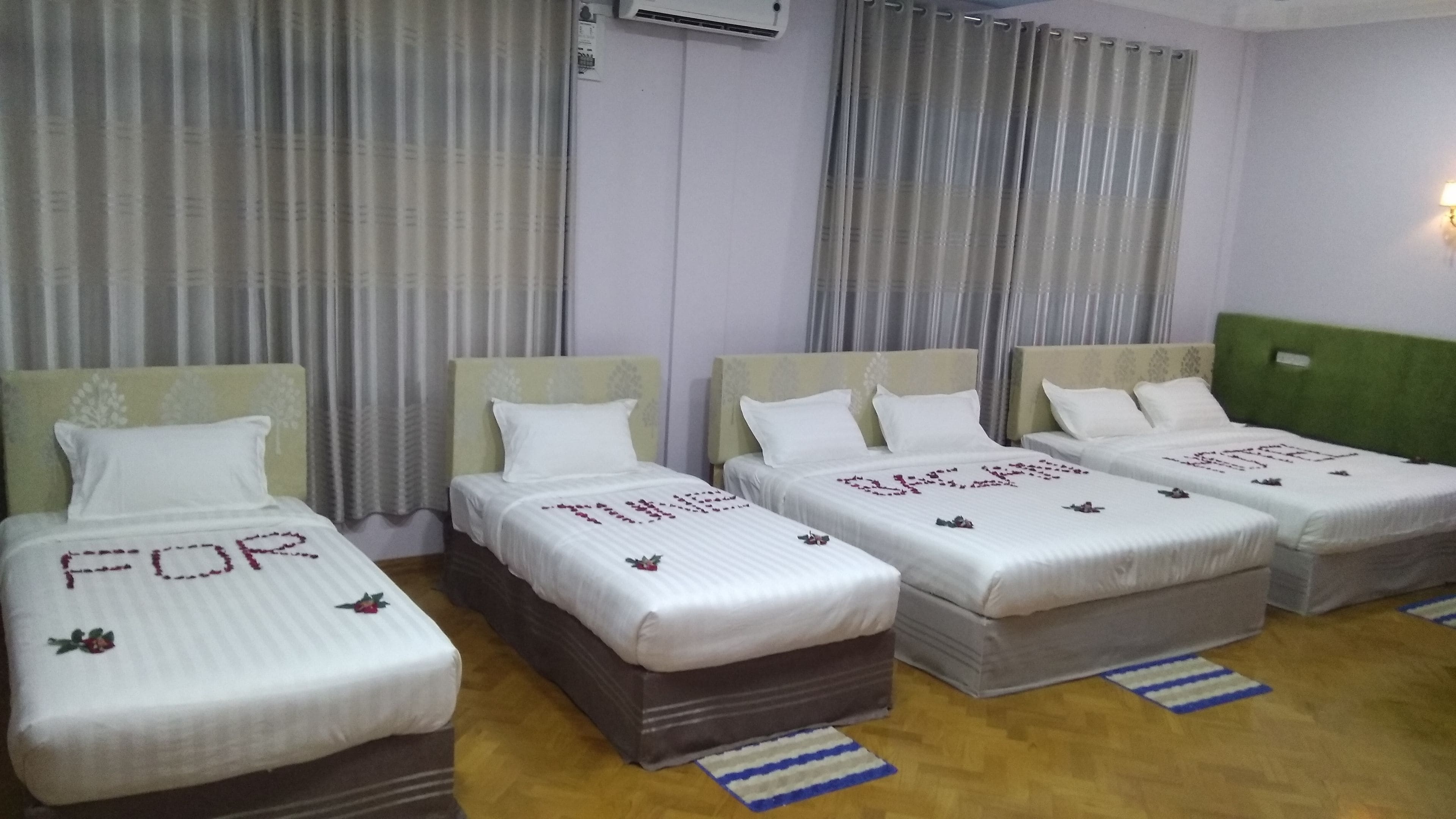 A hotel room with four single beds, each with a white bedspread and a floral design. The room has a wooden floor and grey curtains.