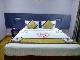 Una cama bien hecha con sábanas blancas, dos almohadas decorativas, un diseño con forma de corazón en la colcha y una mesita de noche con un control remoto.