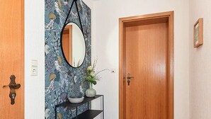 Een gang met een houten deur, een zwart console tafeltje met een vaas, en een spiegel aan de muur.
