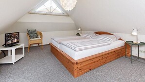 Een slaapkamer met een schuine kap, een bed met gestreept beddengoed, een rieten stoel, een klein tafeltje met een lamp, en een televisie op een witte standaard.