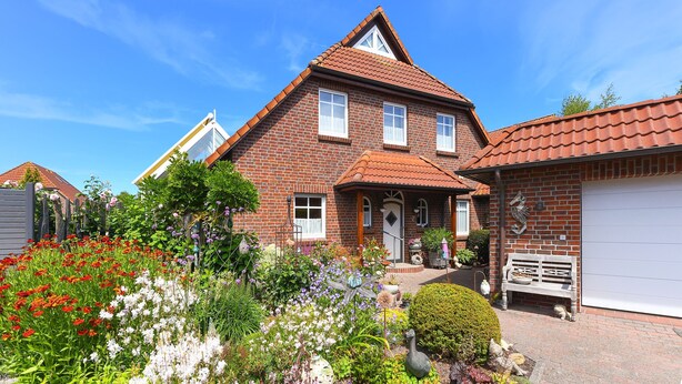 Een twee verdiepingen tellend huis van baksteen met een dak van rode tegels, een veranda en een tuin met verschillende planten en bloemen.