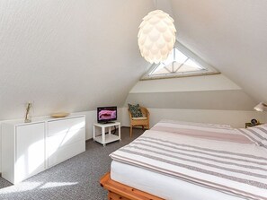 Ein Schlafzimmer mit schrägverdeckter Decke, einem Bett mit gestreifter Bettwäsche, einem Fernseher auf einem kleinen Tisch, einer weißen Kommode und einer Pendelleuchte.