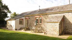 A stone building with a sloped roof, a small patio with a table and chairs, and a grassy yard.