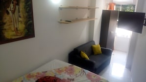 Ein Zimmer mit einem Bett, einem dunkelgrauen Sofa mit gelben Kissen, einem Wand-Fernseher und einem Gemälde an der Wand.