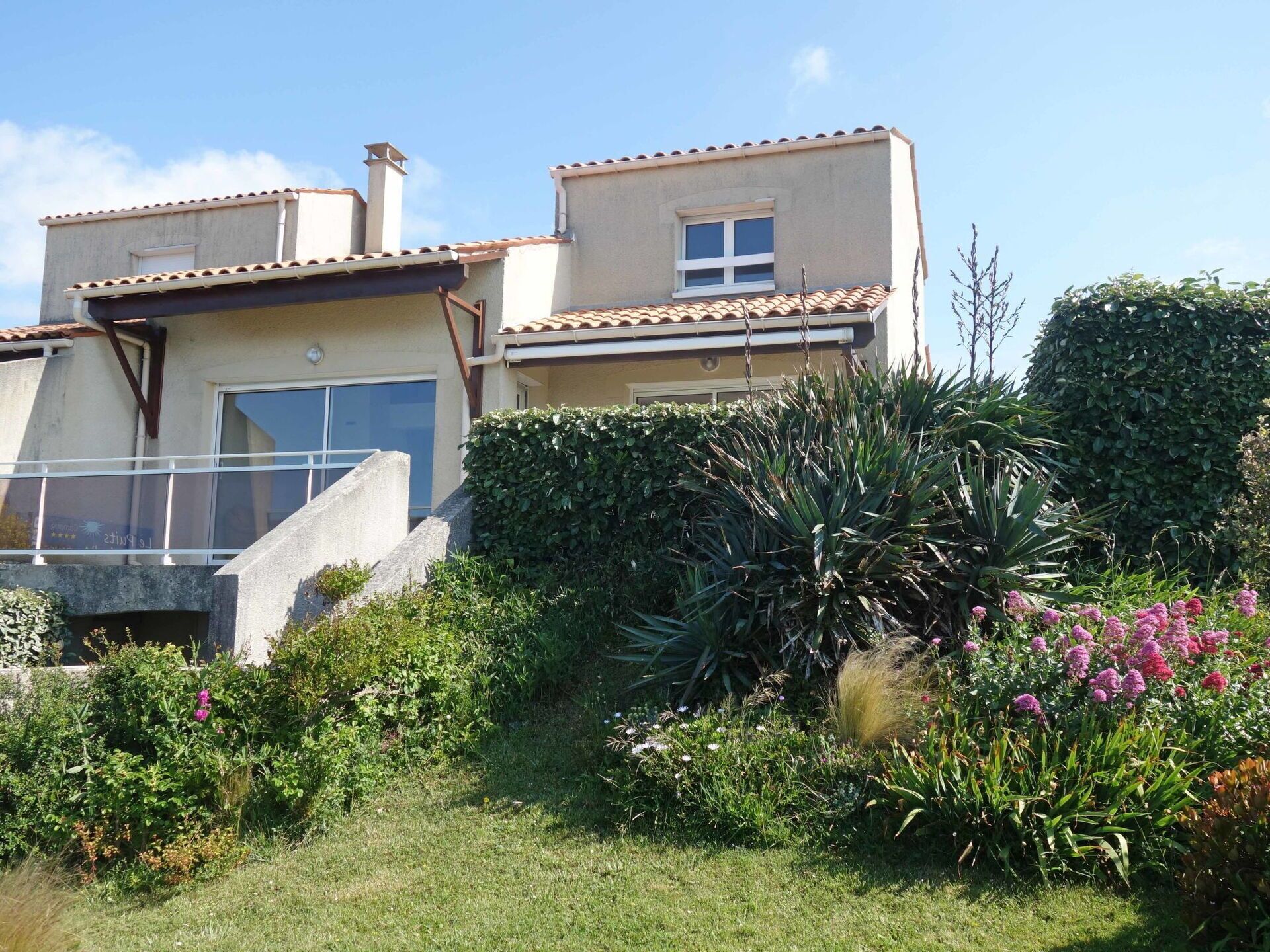 Une maison à deux étages dotée d’un balcon, entourée d’une végétation luxuriante et de plantes en fleurs.