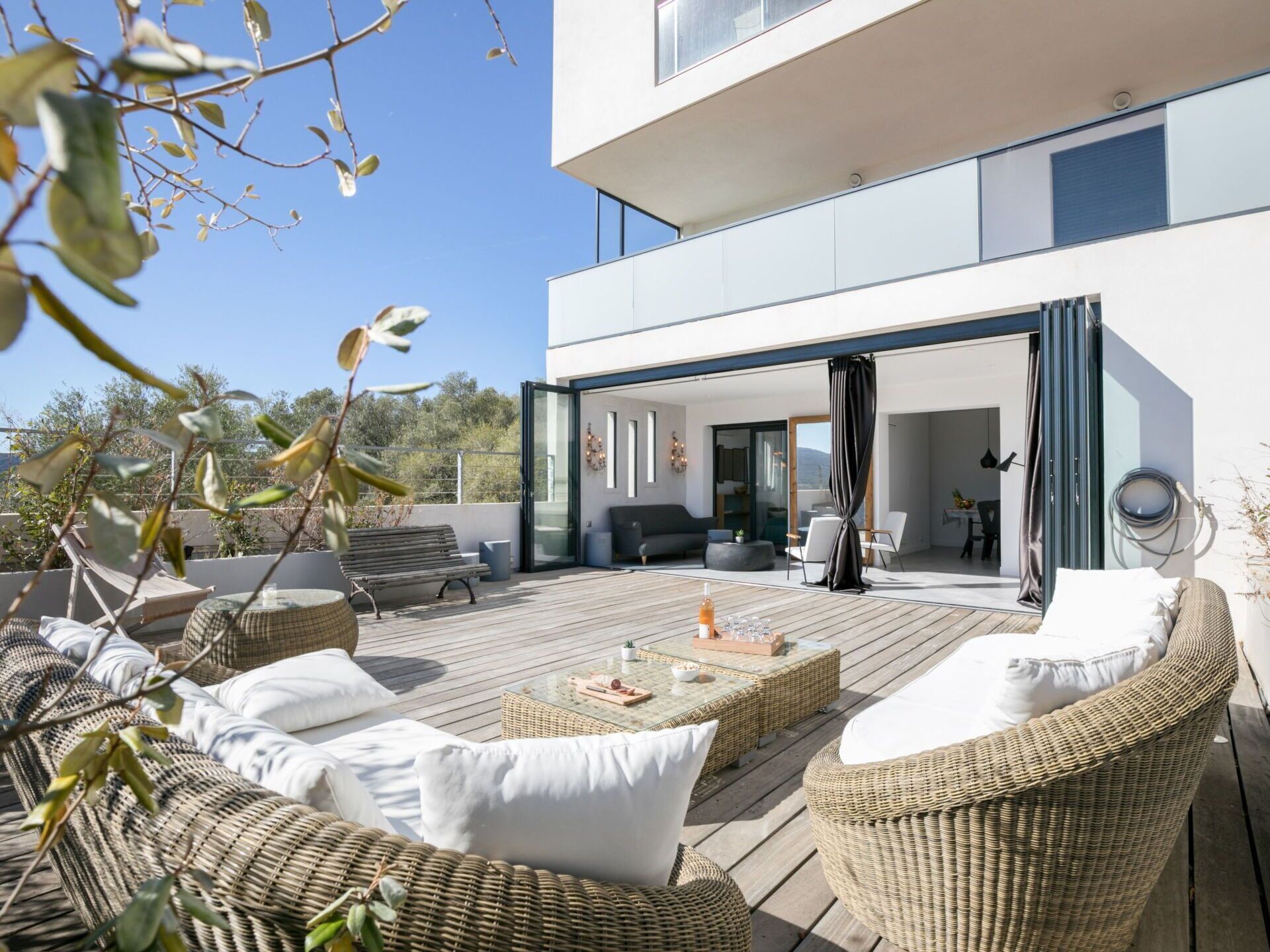 Una terraza moderna al aire libre con mobiliario de mimbre, un ventanal de vidrio que da a un espacio interior y una vista despejada del paisaje circundante.
