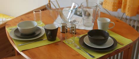 Une table à manger dressée avec des bols, des tasses et des couverts, ainsi qu’une carafe en verre transparent et un thermos.