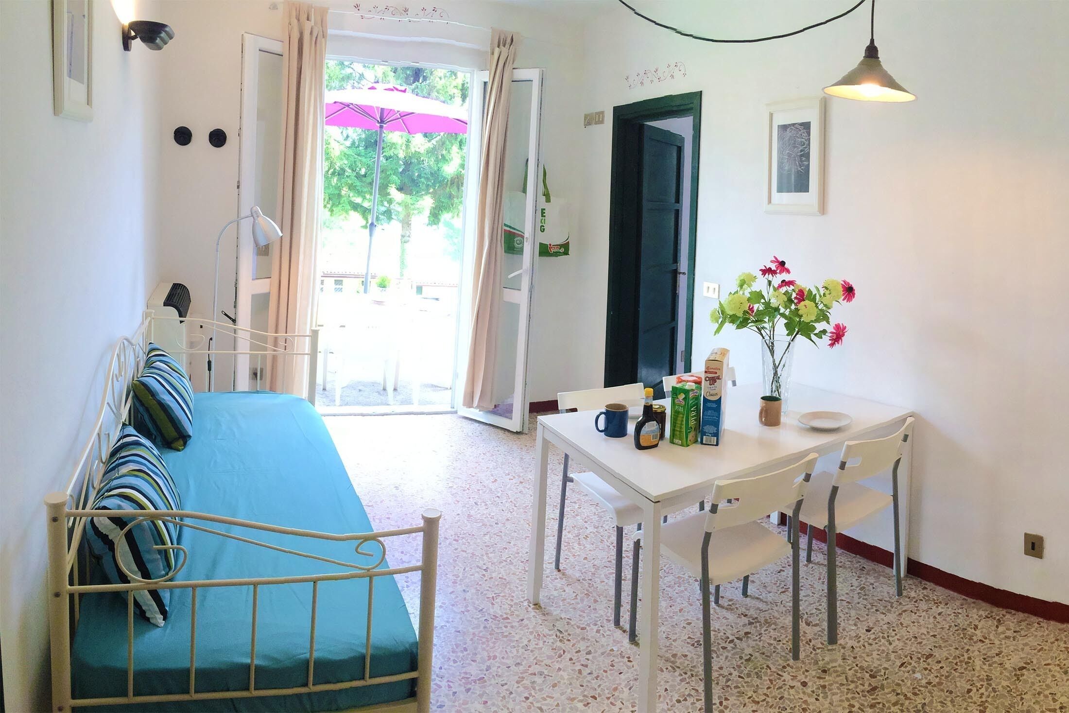 Ett rum med en säng, ett bord med stolar, en vas med blommor och en dörr som leder ut.