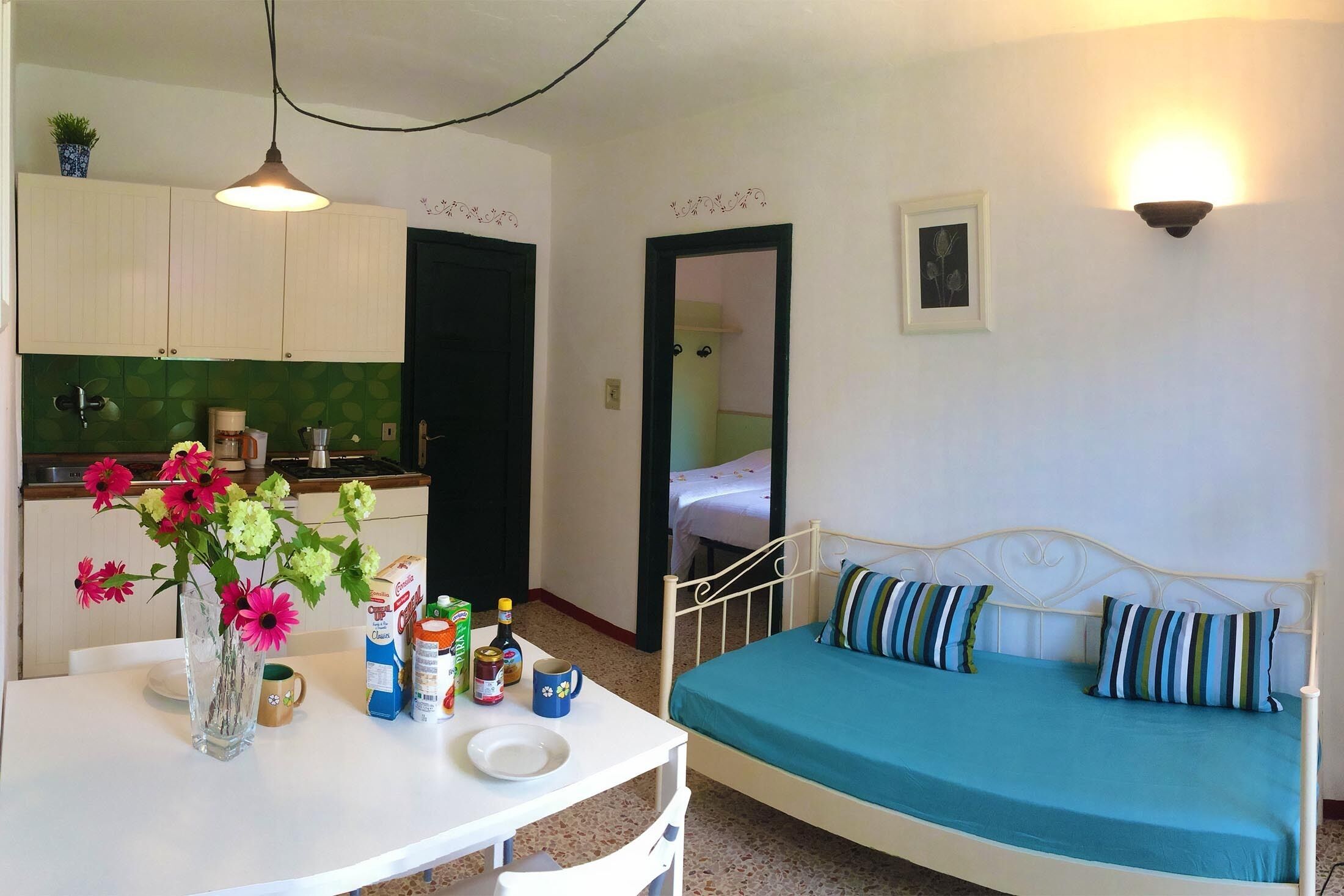A small, clean room with a bed, a table with a vase of flowers, and a kitchenette.