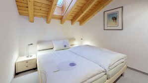 A bedroom with a wooden ceiling, a bed with white bedding, a nightstand, a bedside lamp, and a framed picture on the wall. - Traditional nice apartment house in good location. (Celerina/Schlarigna)