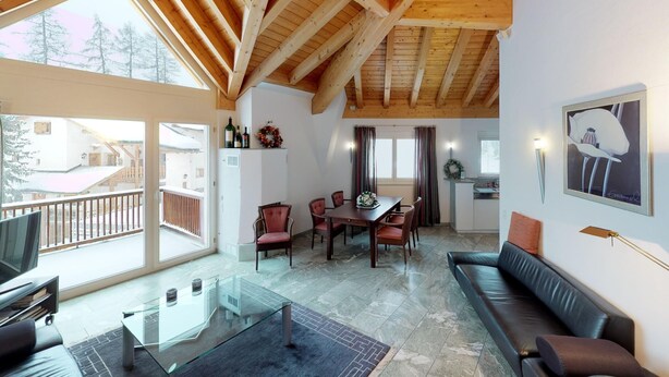 A spacious living area with a dining table, sofa, and a TV. A balcony with a railing is visible, and the room has wooden beams on the ceiling. - Traditional nice apartment house in good location. (Celerina/Schlarigna)