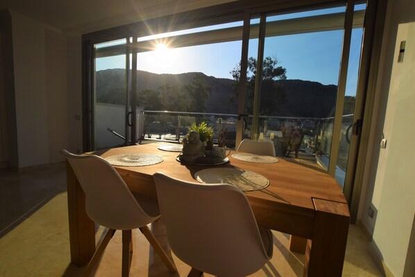 A dining area with a wooden table and chairs, a view of the mountains through large windows, and a balcony with a railing.