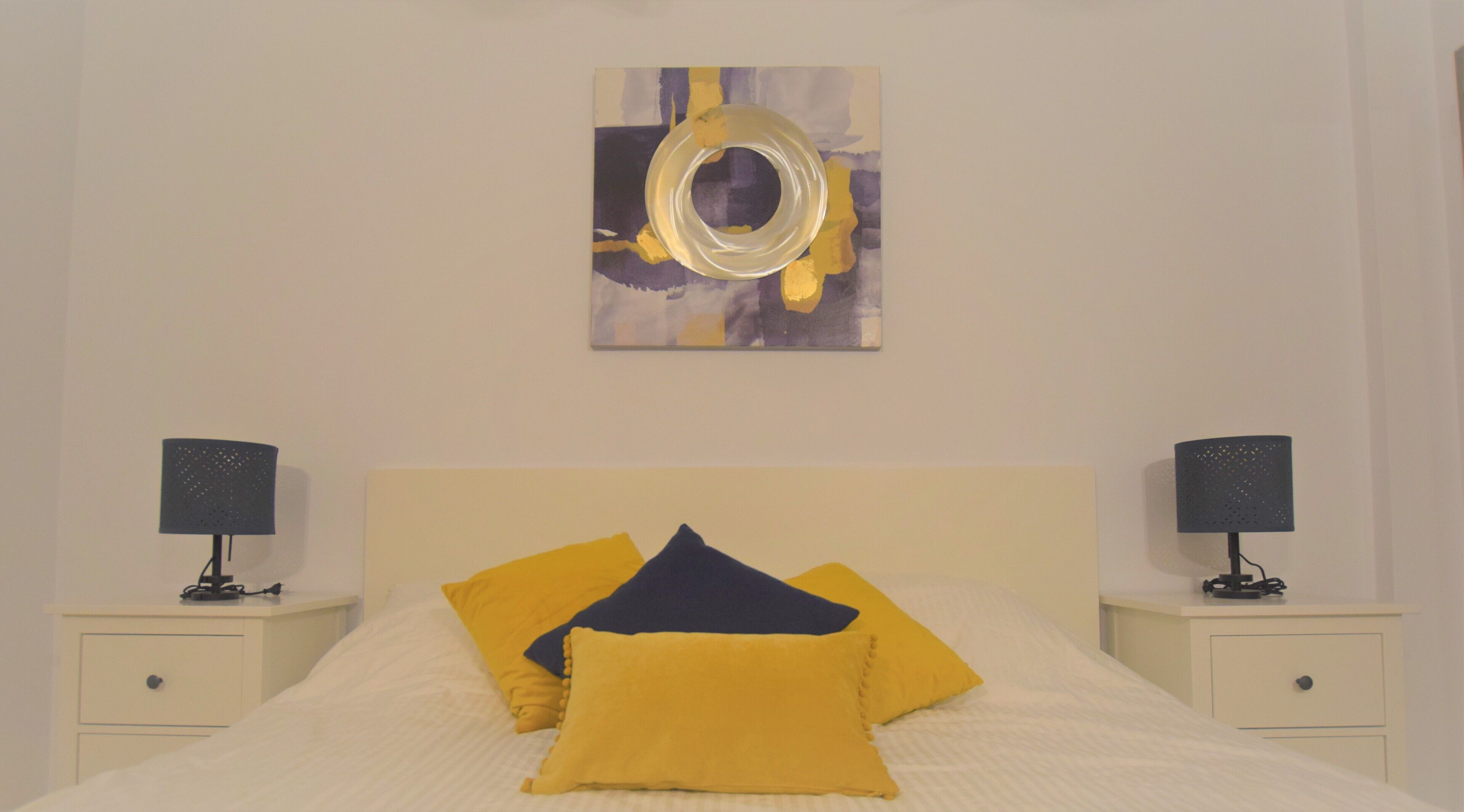 A neatly made bed with yellow and dark blue pillows, two bedside tables with lamps, and a wall-mounted abstract painting.