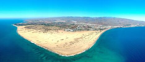 An aerial view of a coastal area with a beach, a town, and a mountain range in the background.