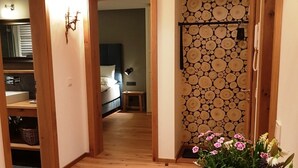 Um corredor com piso de madeira que leva a um quarto com cama e mesa de cabeceira. Há uma planta em vaso com flores sobre um suporte de madeira no corredor.