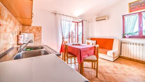 Una cocina compacta con un lavamanos, una mesa de comedor con sillas y un mantel rojo. Hay una ventana con cortinas, un radiador y una planta en maceta en el alféizar.