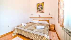 Una cama bien hecha con una colcha a cuadros, toallas enrolladas, una mesita de noche de madera con un libro y una botella, un estante en la pared con vasos, y un cuadro de flores.