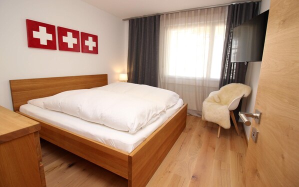 Habitación de hotel con una cama de madera, una silla, un televisor y tres adornos con la bandera suiza roja en la pared.
