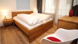 Un lit bien fait, avec du linge de lit blanc, une lampe de chevet, une table de chevet en bois, un bureau en bois, une chaise blanche avec un coussin rouge et un tableau représentant deux patineurs accroché au mur.