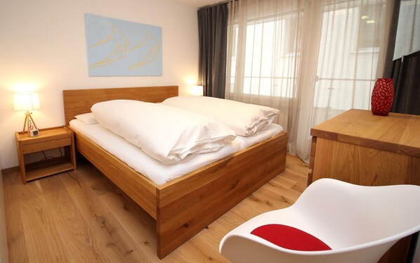 Cama bem arrumada com lençóis brancos, luminária de cabeceira, mesa de cabeceira de madeira, escrivaninha de madeira, cadeira branca com almofada vermelha e um quadro com dois patinadores na parede.