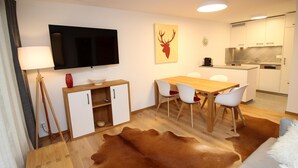 Un salon moderne avec une table à manger en bois, des chaises blanches, un téléviseur à écran plat fixé au mur, un coin cuisine avec des meubles blancs et un tapis en peau de vache.