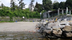 A rocky shoreline with a clear water body, a stone structure, and a covered seating area with chairs. - Studio with a breathtaking view of Lake Maggiore, incl. 1 PP Ged. (Ghiffa Verbania)