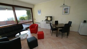 A modern living room with a sofa, red chair, black ottoman, dining table with chairs, a TV, and a refrigerator. - Studio with a breathtaking view of Lake Maggiore, incl. 1 PP Ged. (Ghiffa Verbania)
